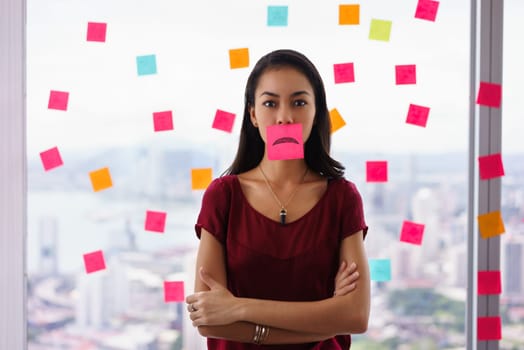 Mixed race woman working in modern office with reminders on skyscraper window. The girl feels stressed, holds a sticking note with sad emoticon on mouth.