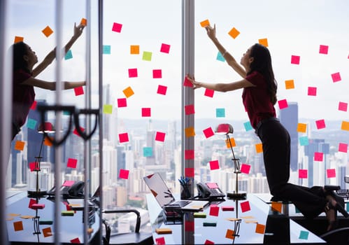 Mixed race secretary working in modern office in skyscraper, sticking adhesive notes with tasks on window. The girl feels stressed and overwhelmed