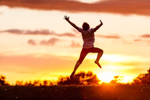 Rear View of a Happy Boy Jumping in the Air with Outstretched Arms and Feet Against Dramatic Sky During Sunset Time.