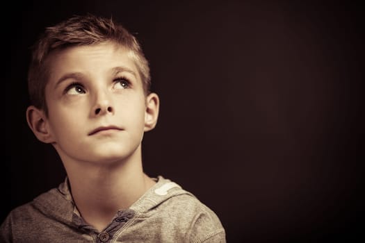 Serious young boy sitting thinking looking up into the air with a pensive expression, head and shoulders over a dark background with copyspace