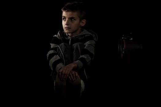 Thoughtful Boy Sitting on the Floor and Holding his Feet with Arms Crossed, Looking Into the Distance Against Black Background.