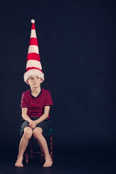 Young boy in a red and white striped dunce cap sitting barefoot in the corner with a dejected expression over a dark background with copyspace
