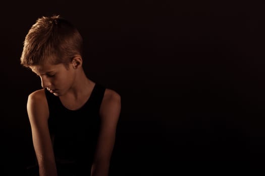 Lonely Slim Boy in Sleeveless Shirt Looking Down Against Dark Background with Copy Space on the Left Side.