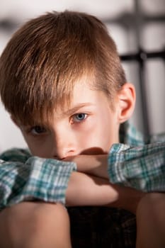 Close up Blond Young Boy with Knees Up and Folded Arms, Looking Straight at the Camera with Sad Facial Expression.