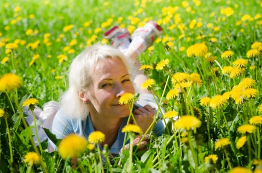 A woman lies in a clearing among the dandelions and sniffs a flower