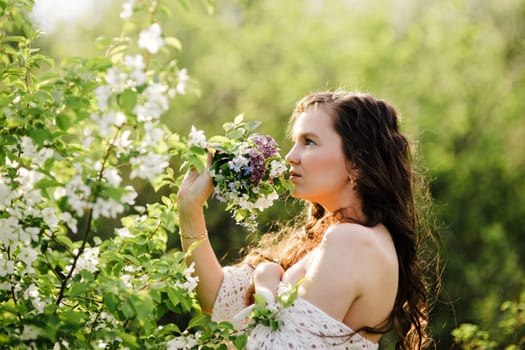 The young girl with a bouquet of wild flowers with naked shoulders in beams of the sun costs near a bush with white flowers