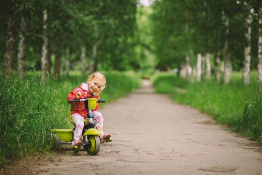 the little girl with white hair in a pink t-shirt goes by small bicycle on the asphalted path in a green grove