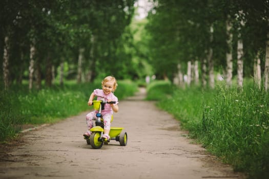 the little girl with white hair in a pink t-shirt goes by small bicycle on the asphalted path in a green grove