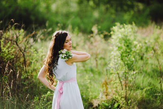 the girl with a bouquet of white flowers in a blue dress with a pink belt poses in the wood