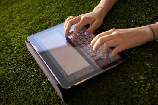 Closeup view of young woman writing on tablet pc on grass, typing an email on digital screen.