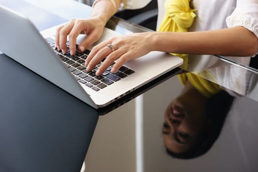 Closeup of hands of young adult woman working as assistant in modern office. The businesswoman writes on laptop pc and smiles. Reflections on table.
