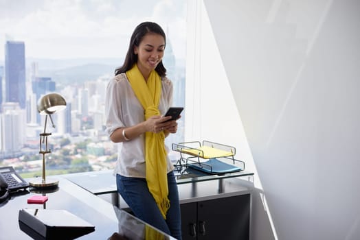 Young multiethnic Chinese Hispanic woman leaning on table in a modern office building, with sight of the city in background. She holds a mobile phone and smiles happy