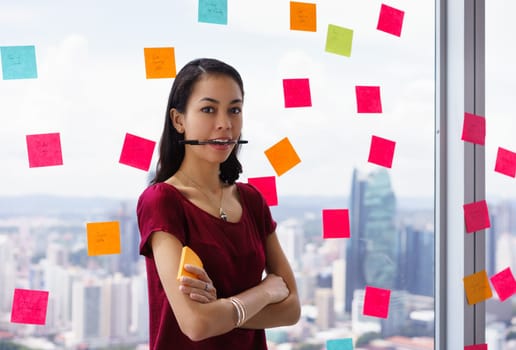 Portrait of proud assistant with pen in mouth. The mixed race woman stands in modern office against a window full of adhesive notes with tasks