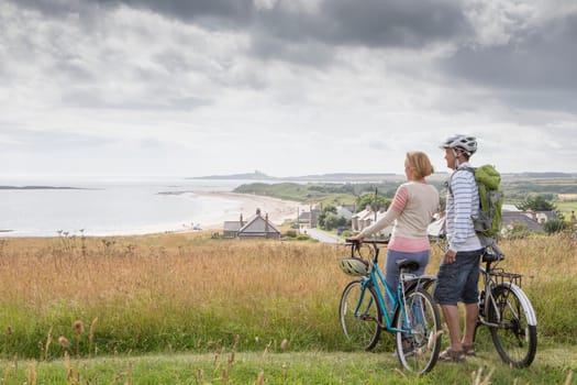 A couple are enjoying the view on the sand dunes. They are holding their bikes, looking at the beach.