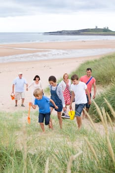 A family are at the beach together, they are all walking up the sand dunes, smiling and talking.