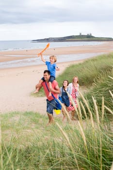 A family of four are walking up the sand dunes toegther, leaving the beach. The little boy is on his dads shoulders laughing.