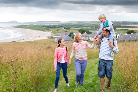 A family of four are walking on the sand dunes together. The son is sitting on his fathers shoulders. They all look happy and are smiling.