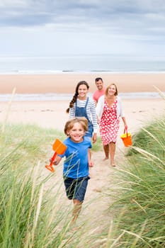 A family of four are walking up the sand dunes leaving the beach. The little boy is running up first smiling at the camera.