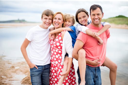 A family of five are stood on the beach together. They are all smiling, looking a the camera.