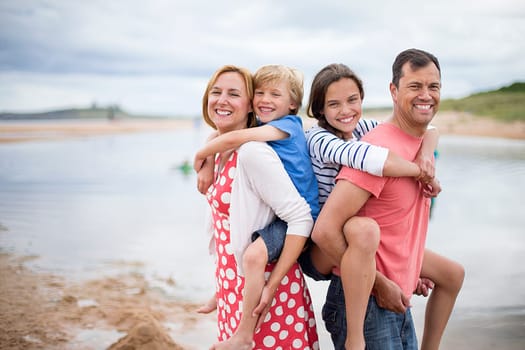 A family of four are on the beach. The parents have the children on thei back, they are all smiling looking at the camera.
