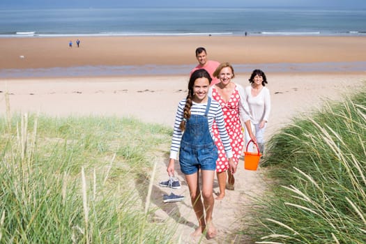 A family of four are walking up the sand dunes leaving the beach. They all look happy and are smiling.