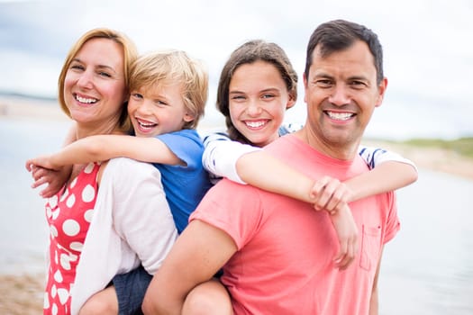 A family of four are at the beach together, the children are on their parents backs. They are all smiling looking at the camera.