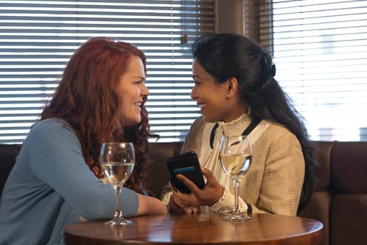 Two women, sitting in a pub looking at a smartphone together. One of the women is transgender. They are both laughing and looking at each other.