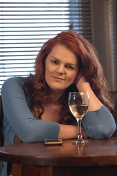 Attractive young woman sitting in a pub with a glass of wine and her smartphone. She is smiling at the camera.
