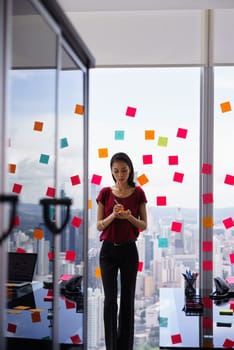 Mixed race secretary working in modern office in skyscraper, writing and sticking adhesive notes with tasks on window.