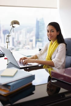 Portrait of mixed race business woman in casual clothing, sitting in modern office. The secretary writes on laptop and smiles at camera. Medium shot