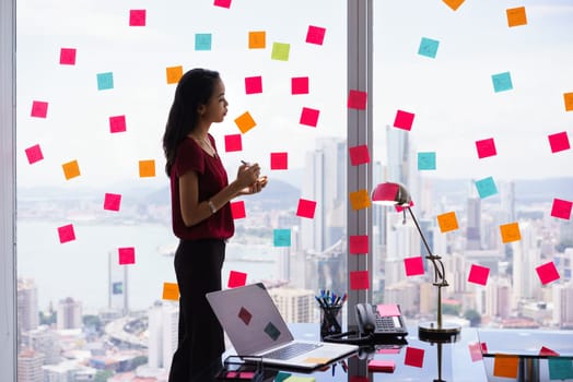 Mixed race secretary working in modern office in skyscraper, writing and sticking adhesive notes with tasks on window.
