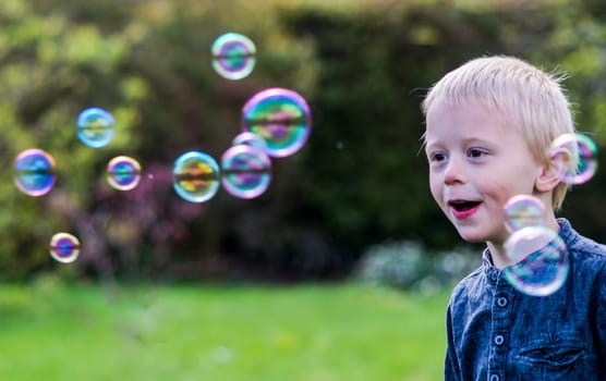 A Little boy blows soap bubbles in the garden on a summer day. He wears a blue shirt and is happy