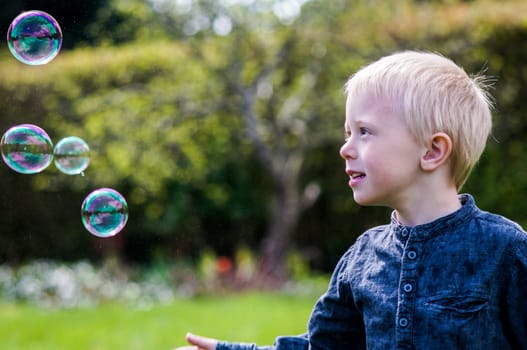 A Little boy blows soap bubbles in the garden on a summer day. He wears a blue shirt and is happy