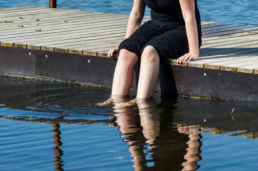young lady sitting on the jetty and dips her feet in the cool lake water on a sunny day