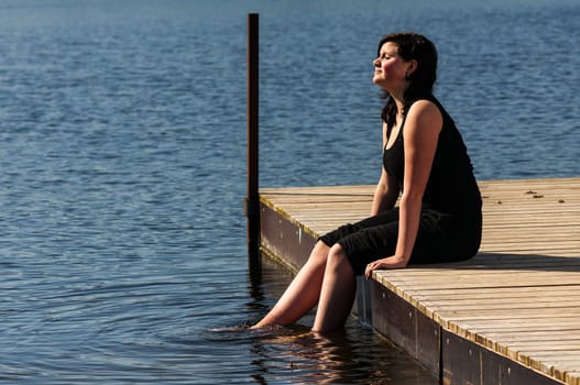 young lady sitting on the jetty and dips her feet in the cool lake water on a sunny day