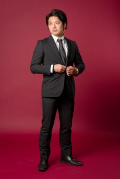 A portrait of a young Japanese man in a business suit standing in a strong pose while buttoning his Jacket on a red background.