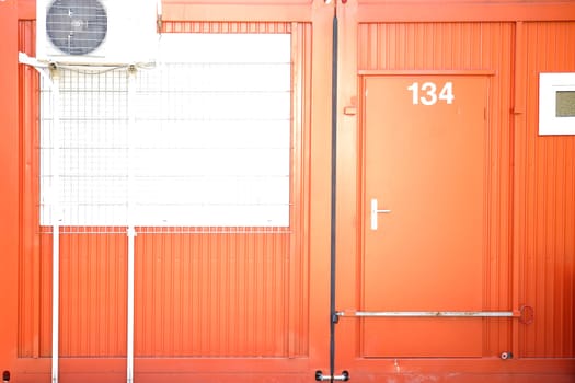 A bright orange-coloured construction container with a locked entrance door and a barred window.