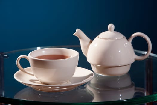 White ceramic cup of tea with saucer and teapot on a glass table with a blue background.