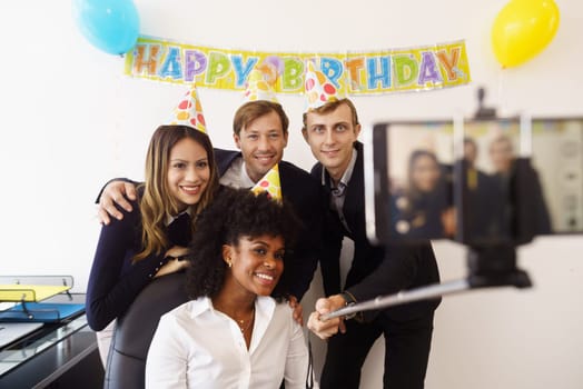 Business woman celebrating her birthday and doing a party with colleagues in her office. A friend holds his phone with a selfie stick and takes pictures of his fellow coworkers smiling. Medium shot