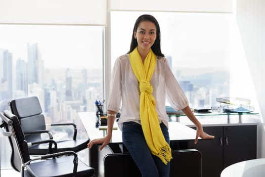 Young multiethnic Chinese Hispanic female entrepreneur leaning on table in a modern office building, with sight of the city. She looks at camera and smiles happy