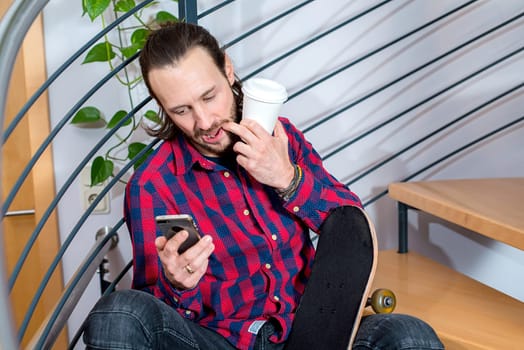 young bearded man in checkered shirt sitting on stairs and using phone