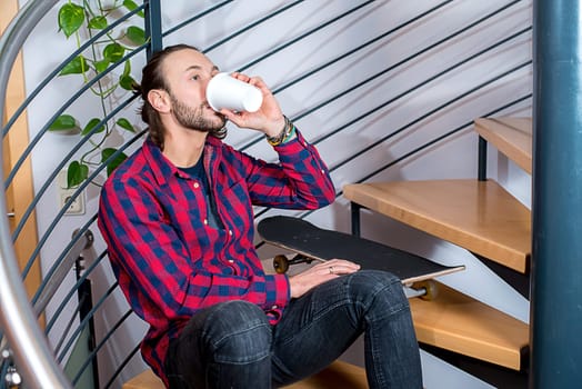 young bearted man in checkered shirt sitting on stairs and drinking coffee