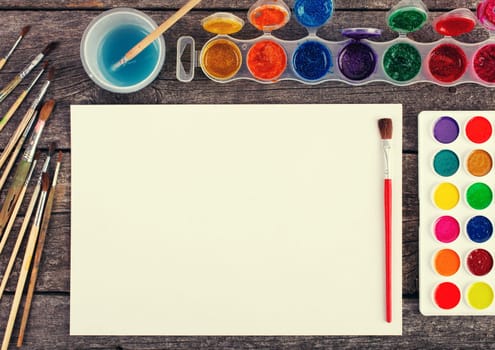 Set of watercolor paints,  brushes for painting and blank white paper sheet of sketchbook on vintage wooden background. Top view.