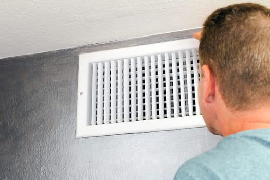 Mature male peering inside an upper wall white grid air duct on a silver wall near a white ceiling. A guy inspecting a heating and cooling air register duct for maintenance.