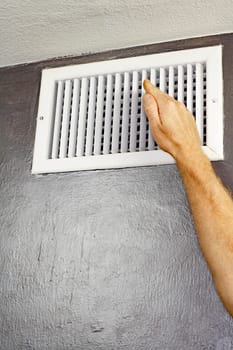 One hand of an adult male raised in front of an upper wall air vent checking for air flow. A man holding a hand up in front of an air vent checking the air temperature..