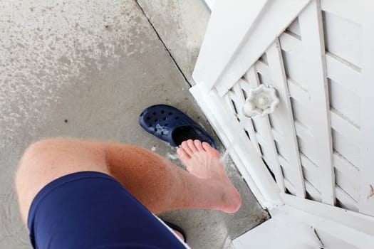 Right leg of a mature male wearing blue sports shorts rinsing sand from his foot after visiting the beach. Outdoor water spigot spraying sand off a man’s right foot outdoors