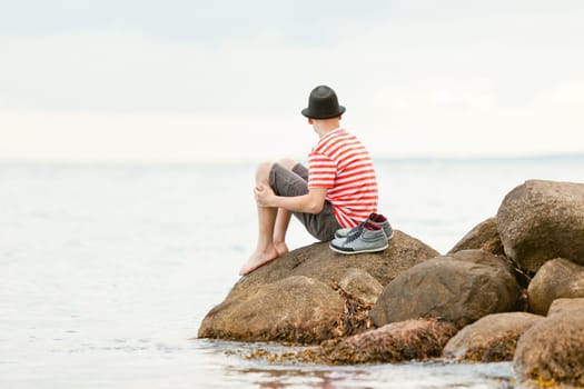 Trendy barefoot young man sitting on a rock at the edge of the ocean staring away from the camera out over the water, with copy space