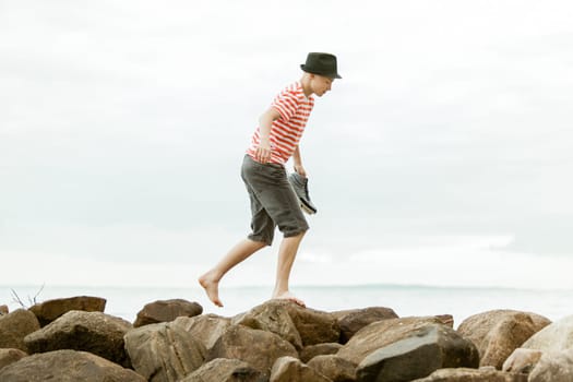 Single teen in black hat and striped shirt holding shoes as he walks barefoot on large rocks near ocean waterfront
