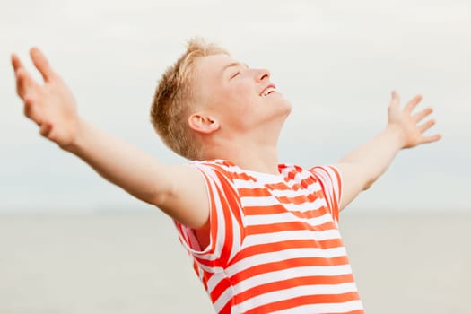 Close up of young short haired teenager in striped shirt with big smile, closed eyes and wide open arms standing near water