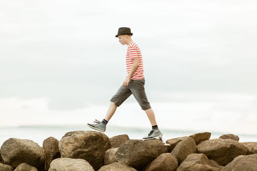 Teenage boy striding across rocks at the seaside in a trendy summer outfit, side view against an ocean backdrop